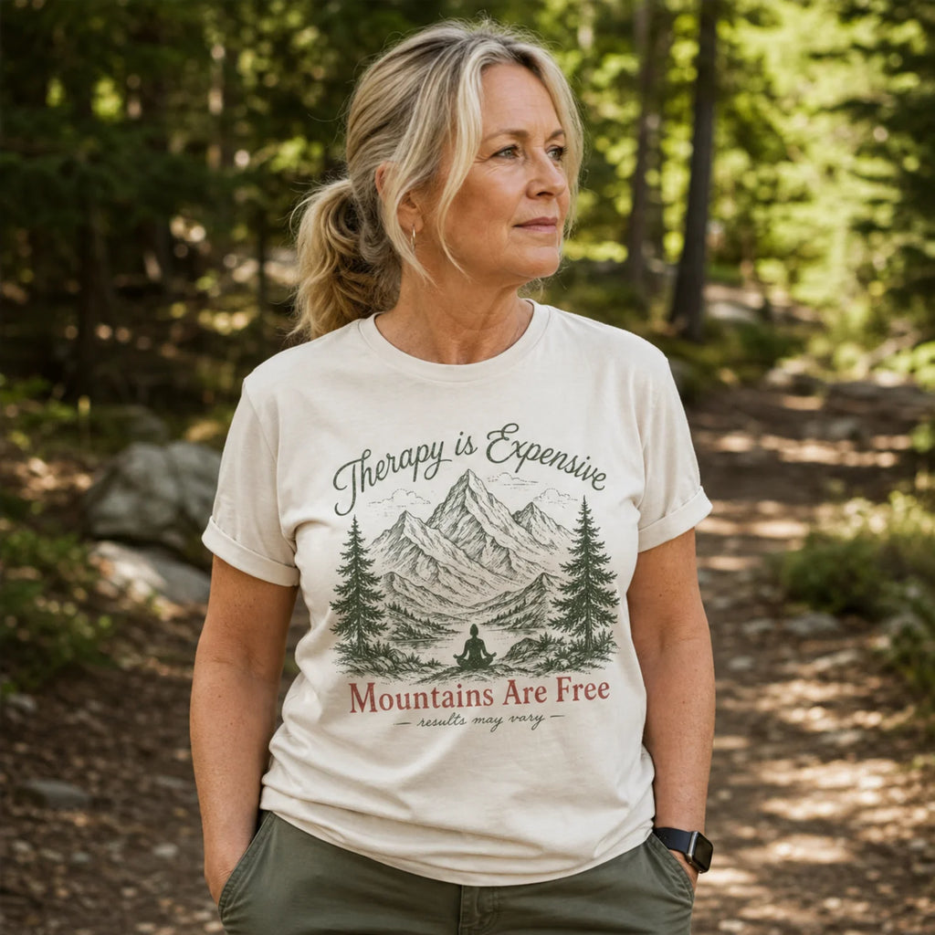 A woman wearing a cream-colored graphic t-shirt with a mountain and pine tree design and the text 'Therapy is Expensive' and 'Mountains Are Free', standing in a sun-dappled forest setting.