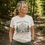 A woman wearing a cream-colored graphic t-shirt with a mountain and pine tree design and the text 'Therapy is Expensive' and 'Mountains Are Free', standing in a sun-dappled forest setting.