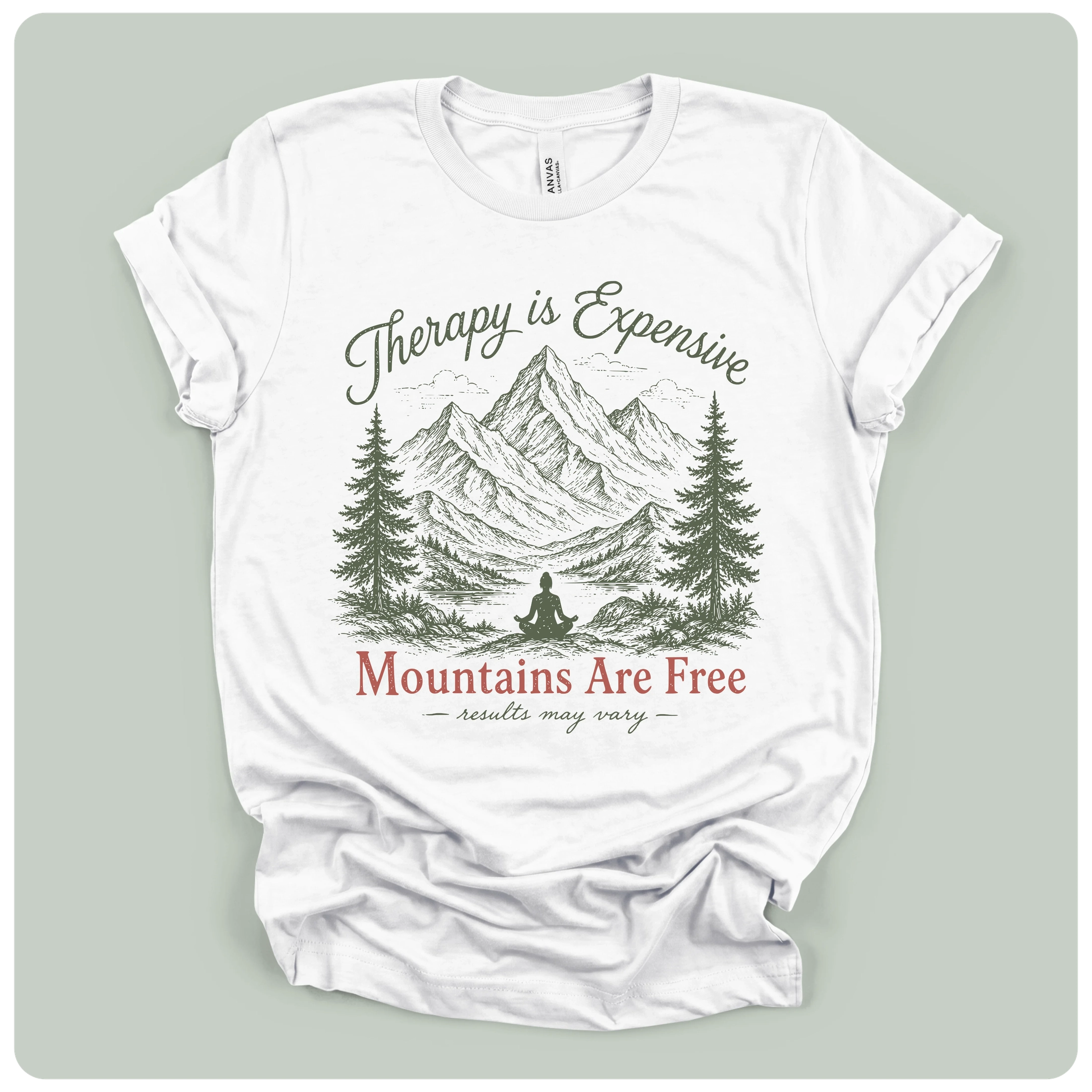A white short-sleeve graphic T-shirt laid flat on a light sage green background. The shirt features a printed design of mountains, pine trees, and a meditating figure, with the text 'Therapy is Expensive' in dark green script and 'Mountains Are Free' in red serif font.