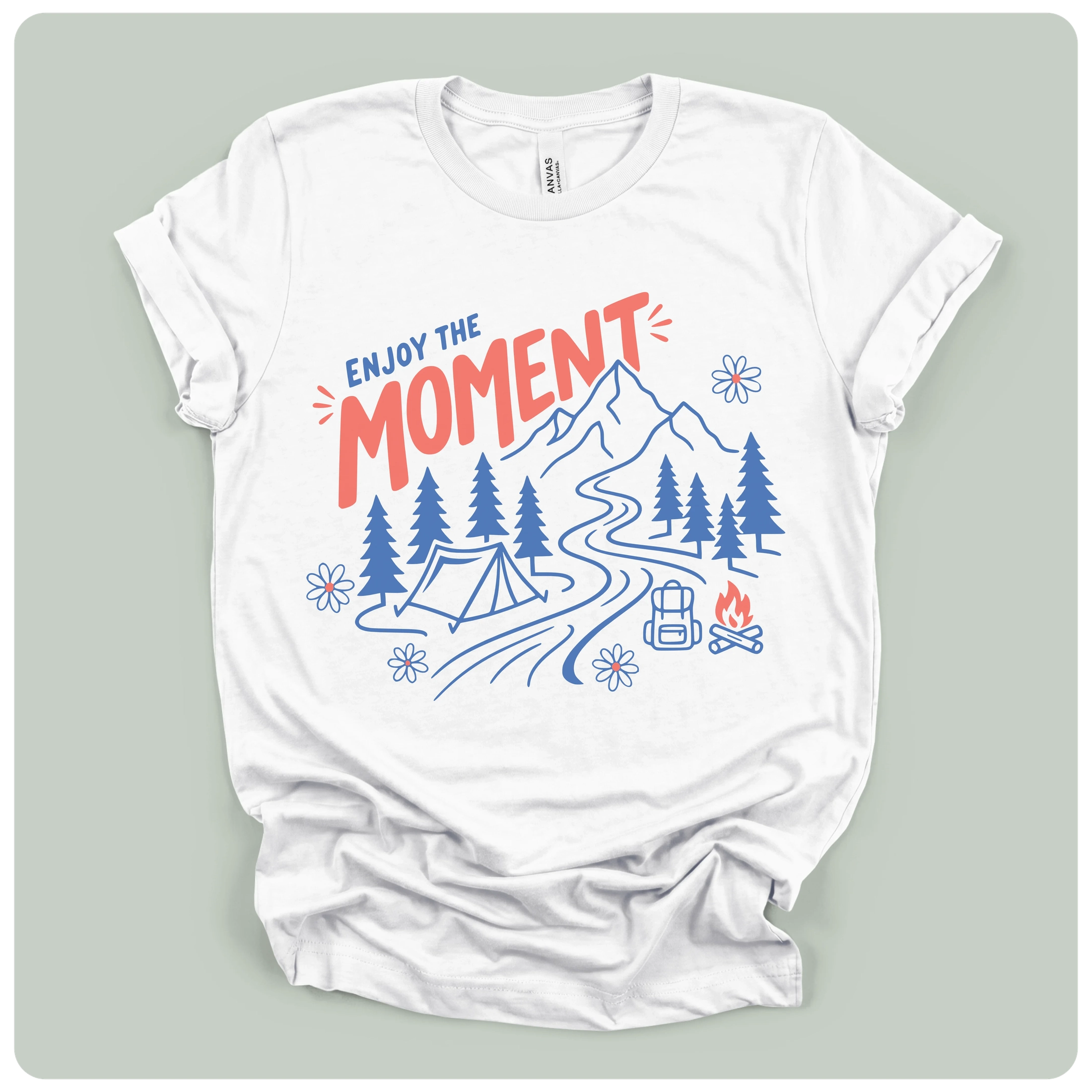 A white short-sleeve t-shirt with a colorful outdoor camping graphic on the front, laid flat on a light sage green background. The graphic features the text 'ENJOY THE MOMENT' in red and blue, with blue line art of mountains, pine trees, a tent, a campsite path, a backpack, and a campfire.