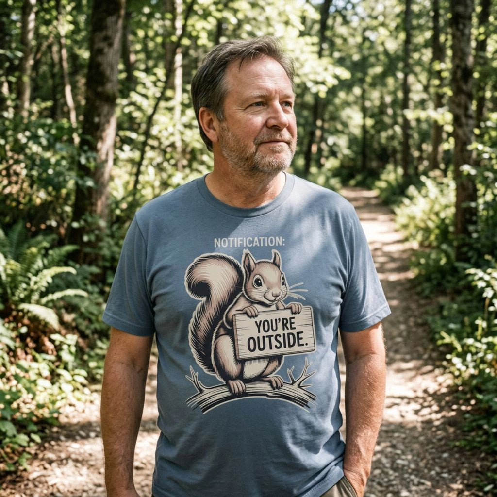 A man standing in a sunlit forest wearing a slate blue graphic t-shirt with a cartoon squirrel holding a sign that reads 'YOU'RE OUTSIDE'.