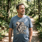 A man standing in a sunlit forest wearing a slate blue graphic t-shirt with a cartoon squirrel holding a sign that reads 'YOU'RE OUTSIDE'.
