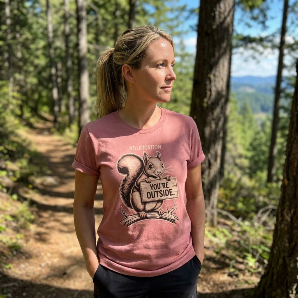 A woman standing on a forest trail wearing a dusty rose pink graphic t-shirt with a squirrel holding a sign that says 'YOU'RE OUTSIDE'.