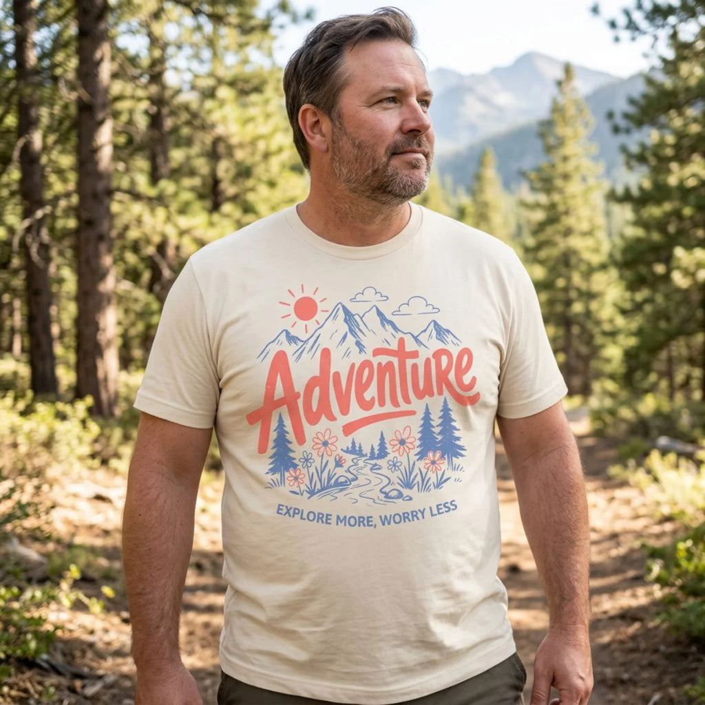 A man wearing a cream-colored graphic t-shirt with a red and blue mountain design that reads 'Adventure Explore More, Worry Less', standing in a forest with mountains in the background.