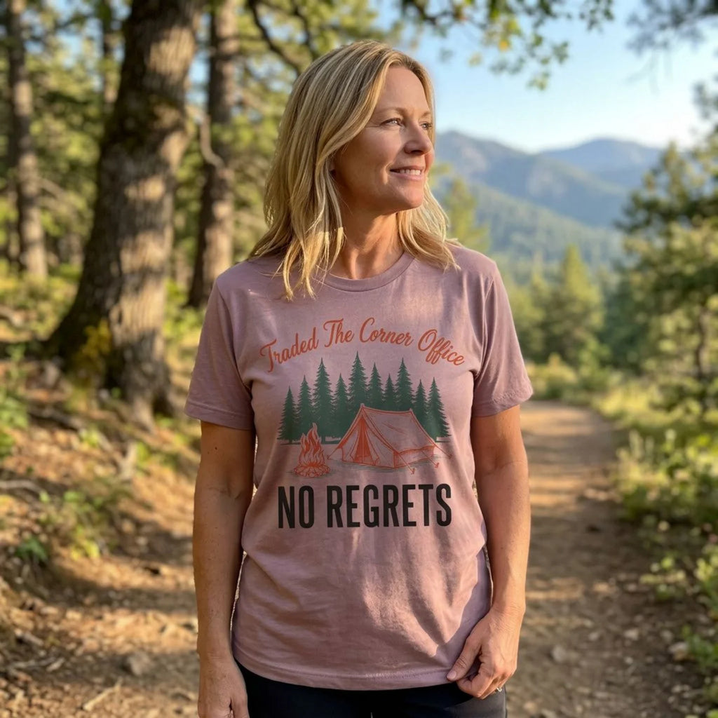 A woman standing on a forest path wearing a dusty pink graphic t-shirt with a camping design and the text 'Traded The Corner Office NO REGRETS'.