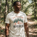 A man standing in a sun-dappled forest wearing a cream-colored graphic t-shirt with a camping design and the text 'Traded The Corner Office NO REGRETS'.