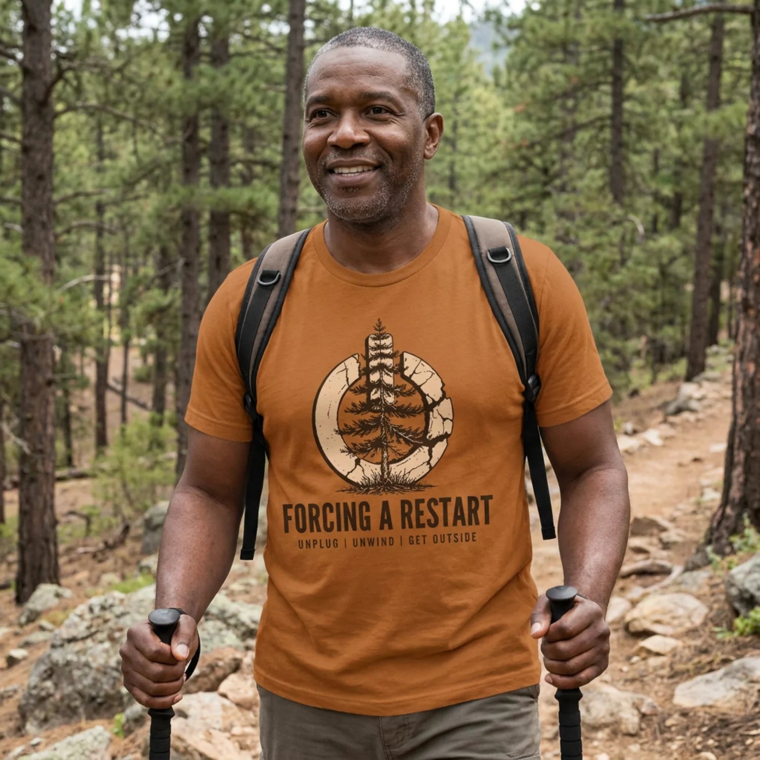 A smiling man hiking in a pine forest wearing an orange graphic t-shirt with a tree illustration and the text 'FORCING A RESTART'.