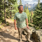 A man standing on a forest hiking trail wearing a sage green graphic t-shirt that reads 'Therapy is Out there Mountains Are Free' with a mountain illustration, paired with khaki pants and hiking boots, with mountains and pine trees in the background.