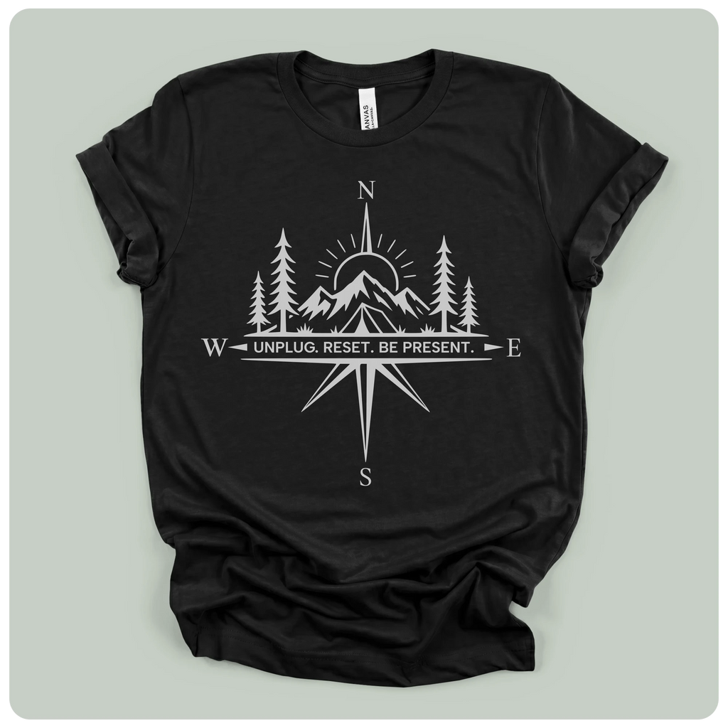 A black short-sleeve crewneck t-shirt with a white graphic print of a compass rose, mountains, pine trees, and a sun, with the text 'W UNPLUG. RESET.