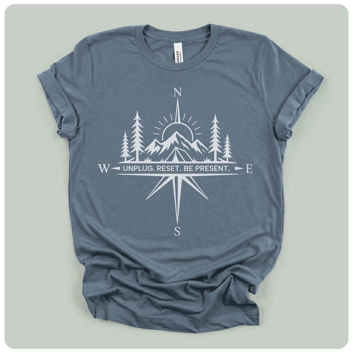 A slate blue crewneck t-shirt with a white graphic print of a compass rose, mountains, pine trees, and a sun, with the text 'W UNPLUG. RESET.