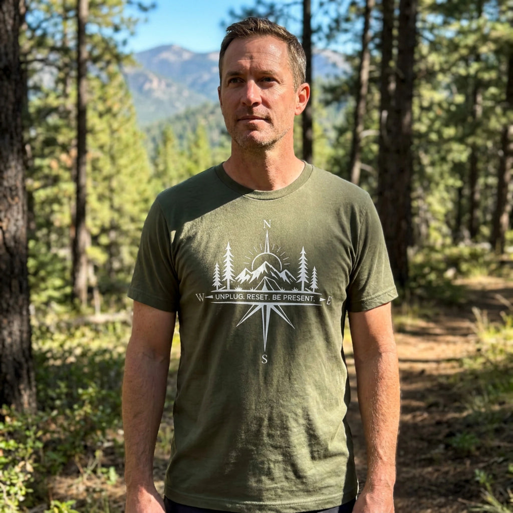 A man wearing an olive green t-shirt with a white compass and mountain graphic on the chest, standing in a forest with mountains in the background.