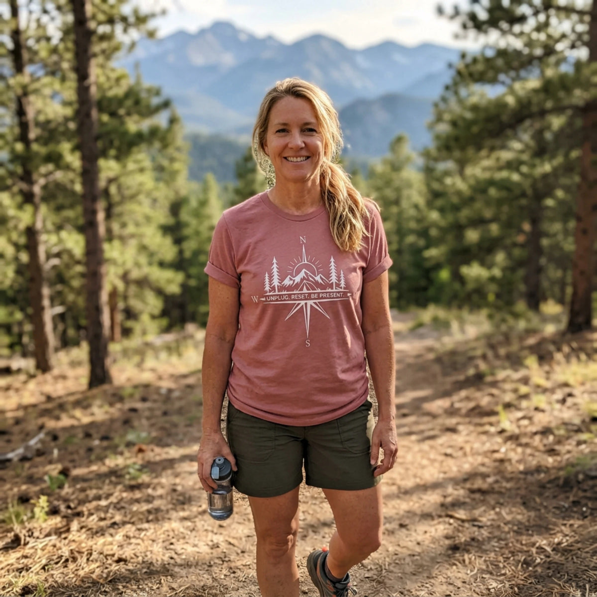A woman hiking on a forest trail wearing a dusty rose t-shirt with a white mountain graphic and the text 'THE PEAK IS NOT THE END' and dark olive green shorts, with mountains in the background.
