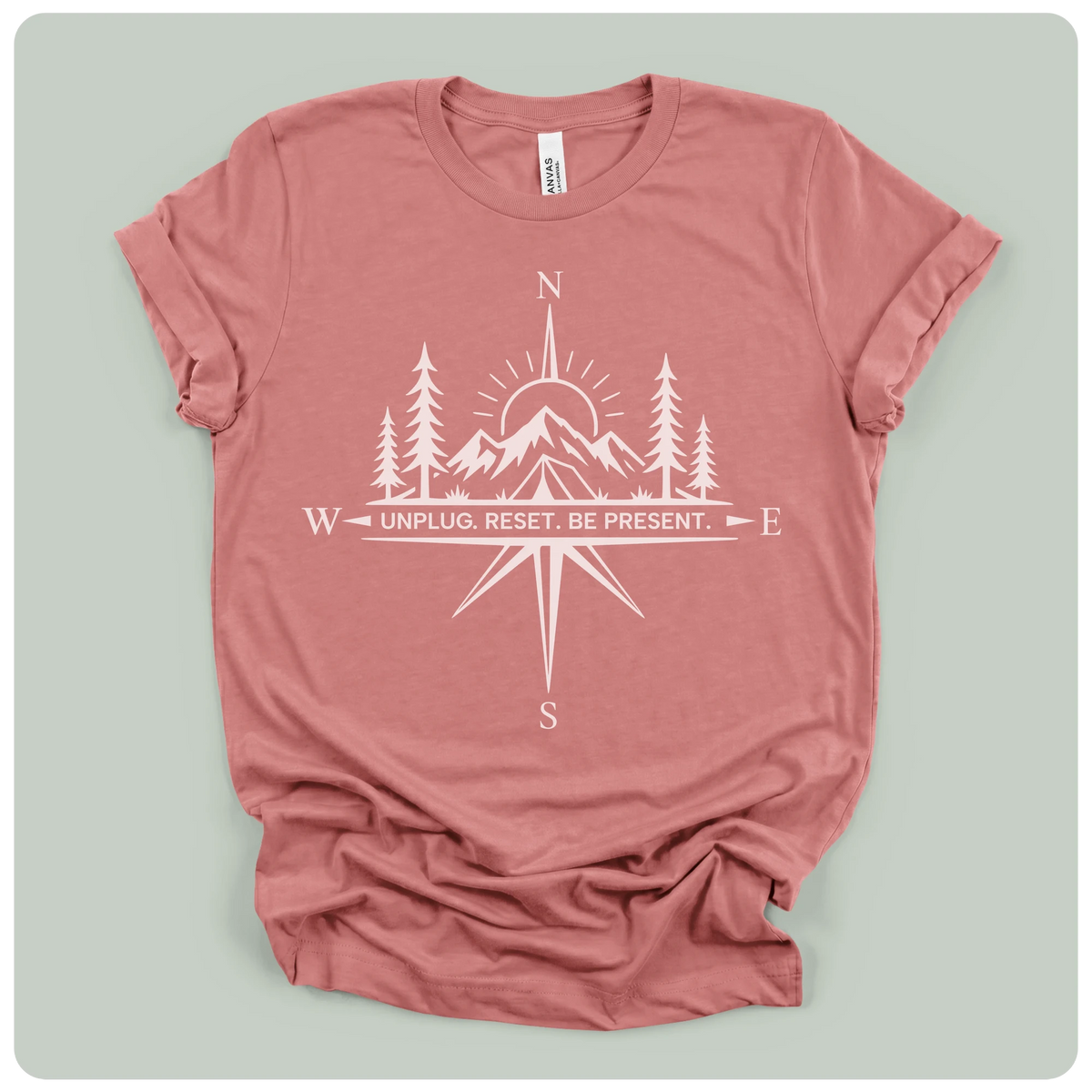 A dusty rose short-sleeve t-shirt with a white graphic print of a compass rose, mountains, pine trees, and a sun, featuring the text 'UNPLUG. RESET.