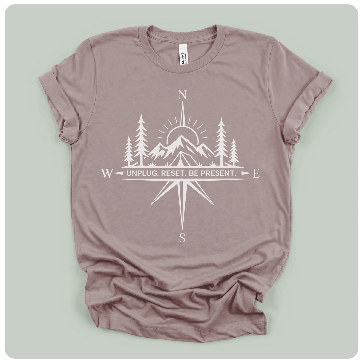A dusty mauve crewneck t-shirt with a white graphic print of a compass rose, mountains, pine trees, and a sun, with the text 'UNPLUG. RESET.