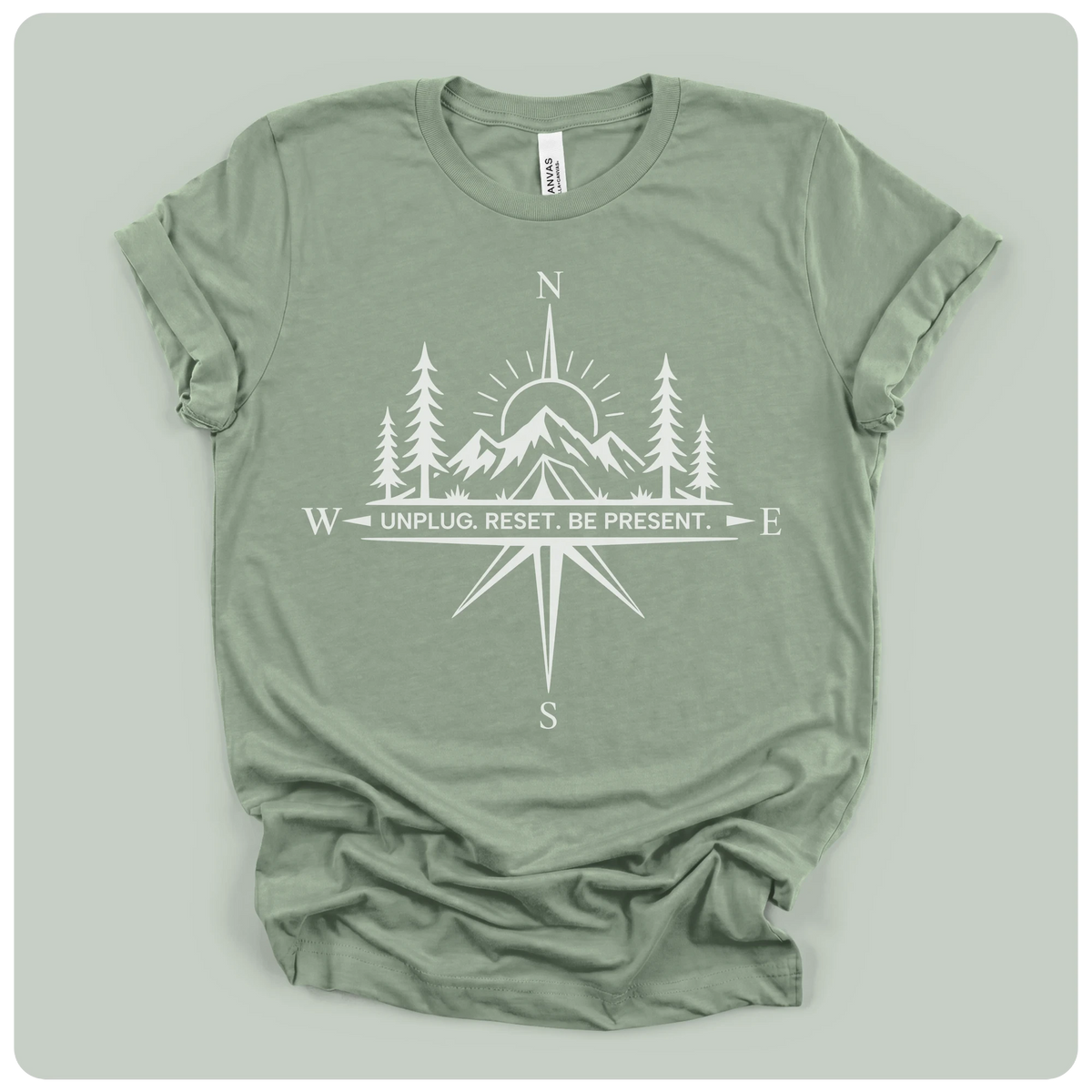 An olive green short-sleeve t-shirt with a white graphic print of a compass rose, mountains, pine trees, and a sun, featuring the text 'UNPLUG. RESET.