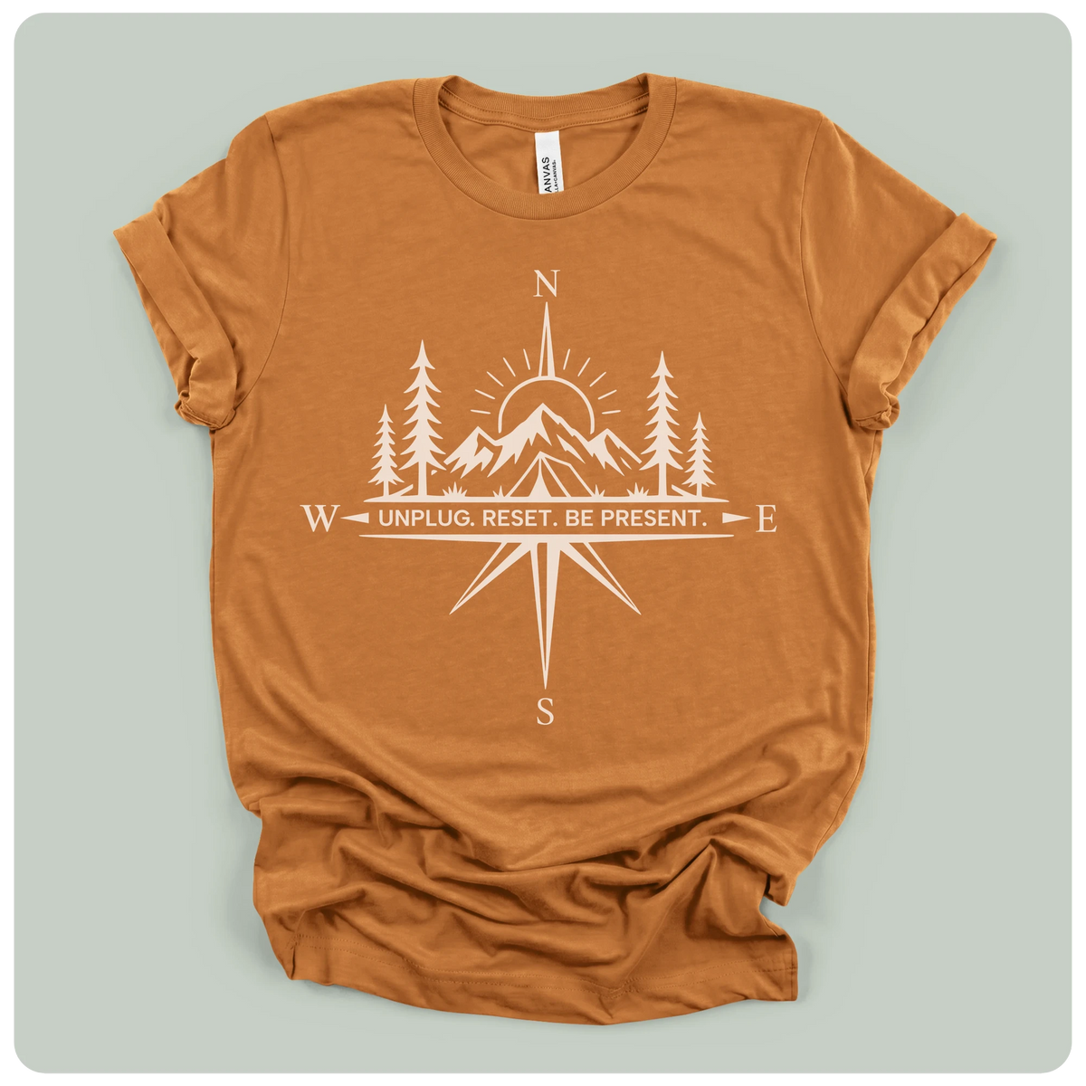 A rust-orange crewneck t-shirt with a white graphic print of a compass rose, mountains, pine trees, and a sun, featuring the text 'W - UNPLUG. RESET.